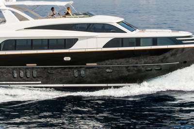 A sleek, dark-hulled yacht cruises through choppy blue water, with two people visible on its upper deck against a hazy sky.
