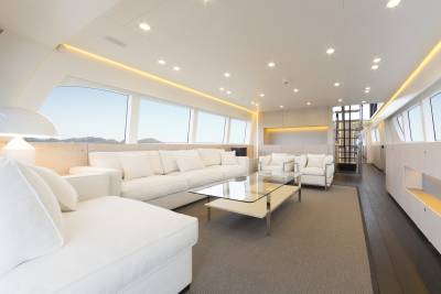 A spacious, modern white living area with plush sofas and a glass coffee table in the foreground is set against a backdrop of large windows offering views of a bright, outdoor landscape.
