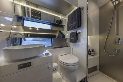 A modern bathroom with a sleek white sink in the foreground and a shower and toilet in the background, all set against a backdrop of reflective surfaces and a window overlooking the water.