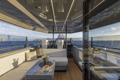 A luxurious yacht's outdoor seating area with a table and champagne in the foreground overlooks a calm sea and rocky islands in the background.