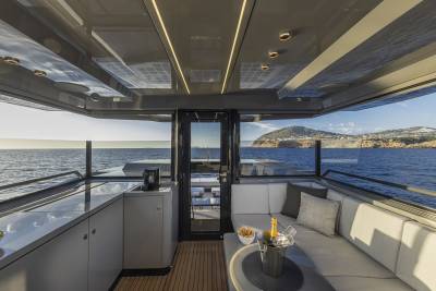 A modern yacht's interior lounge with a wet bar in the foreground and a scenic ocean view with a hilly coastline in the background.
