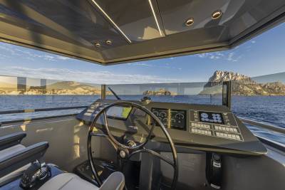 A modern yacht's helm with a large steering wheel and digital displays is positioned in the foreground, overlooking a calm sea and a rocky island in the background under a clear sky.
