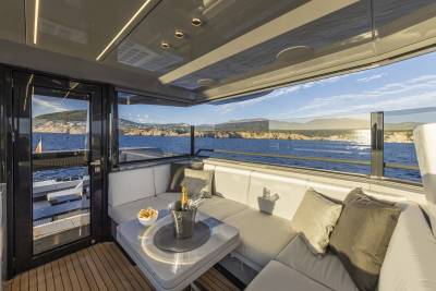 A luxurious yacht's outdoor seating area with a table holding champagne and snacks is set against a backdrop of a calm blue sea and a distant, sun-drenched coastline.