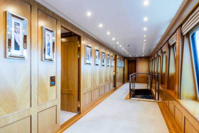 A long, well-lit hallway with framed portraits on the left and windows on the right leads to a staircase descending into darkness.