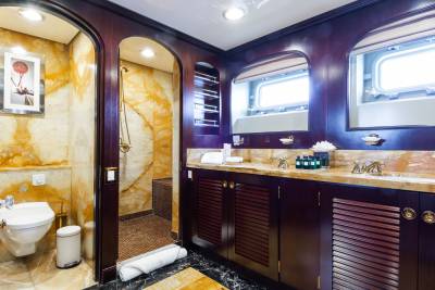 A luxurious bathroom with a marble-tiled toilet area in the foreground and a double vanity with large windows in the background.