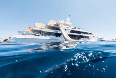 A large white yacht with a slide is anchored in clear blue water, with a rocky coastline visible in the distance under a bright sky.