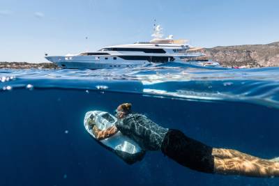 A person glides underwater with a sea scooter in the foreground, while a large white yacht is anchored in the background near a sun-drenched coastline.