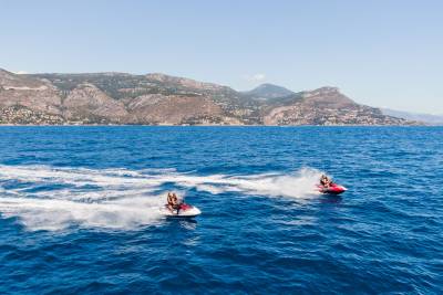 Two jet skis race across the bright blue ocean, leaving white wakes, with a lush, mountainous coastline in the distance under a clear sky.