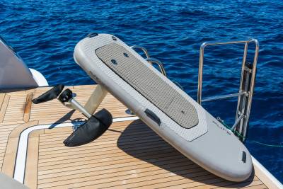 A gray inflatable hydrofoil board rests on a wooden deck in the foreground, with the deep blue ocean visible in the background.