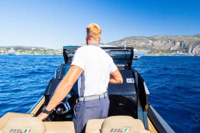 A man with blonde hair steers a boat on a bright blue sea, with a coastline and other boats visible in the distance under a clear sky.