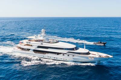 A large white yacht cruises through the blue ocean, with a smaller inflatable boat trailing behind it.