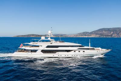 A large white yacht cruises through the blue ocean with a hilly coastline in the background.