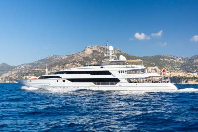 A large white yacht cruises on blue water in the foreground, with a mountainous coastline dotted with buildings in the background under a clear blue sky.