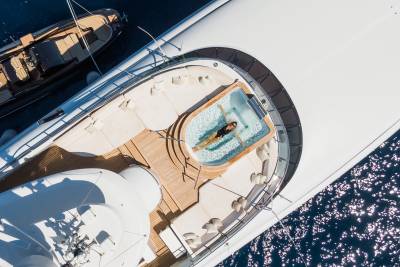 A woman relaxes in a jacuzzi on the deck of a large yacht, with a smaller boat docked nearby and sparkling blue water in the background.