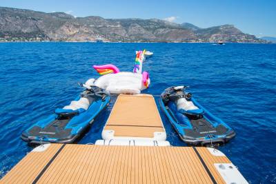 Two jet skis flank a large inflatable unicorn floating on the bright blue sea, with a mountainous coastline visible in the distance.