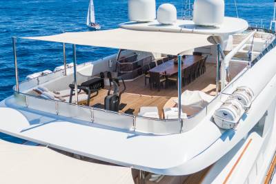 A large yacht's deck features a dining area and exercise equipment in the foreground, with a sailboat on the blue ocean in the background.