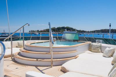 A circular jacuzzi with a waterfall feature is in the foreground, while the background shows a calm blue sea dotted with boats and a lush green coastline under a clear sky.