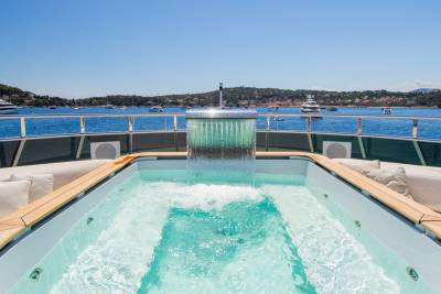 A bubbling jacuzzi on a yacht's deck overlooks a calm blue bay dotted with boats and a distant, tree-lined shore under a clear sky.