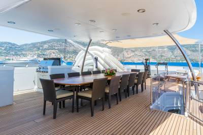 A long dining table with chairs is set on a yacht's deck in the foreground, with a scenic coastal town and mountains visible in the background.