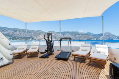 A sun-drenched deck with lounge chairs and exercise equipment faces a picturesque coastline under a clear blue sky.