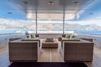 Wicker sofas and a coffee table furnish the wooden deck of a yacht, with a scenic view of the ocean and distant hills in the background.