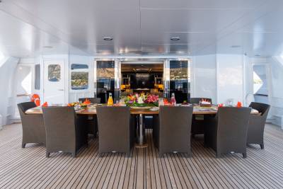 A beautifully set dining table with floral arrangements and drinks sits on a wooden deck, with a view of a luxurious interior and distant hills through open glass doors.