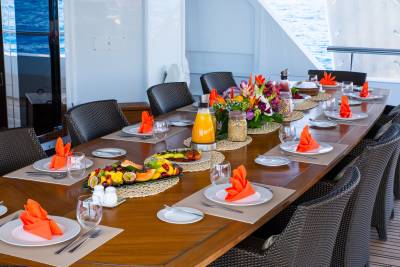 A long wooden table set with orange napkins, fruit platters, and a floral centerpiece is arranged on a yacht deck with the ocean visible in the background.