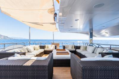 A spacious yacht deck with comfortable seating and a fire pit in the foreground is set against a backdrop of the sparkling blue ocean and distant hills under a clear sky.