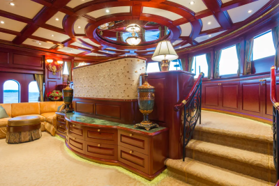 A luxurious yacht interior features a plush cream carpet and ornate wooden cabinetry in the foreground, while the background reveals large windows offering a glimpse of the ocean and a grand, circular ceiling.