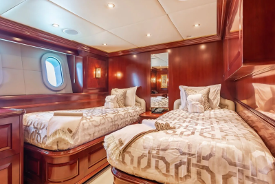 Two neatly made twin beds with patterned bedding fill the foreground, while the background reveals a luxurious cabin with dark wood paneling and oval windows.