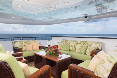 A luxurious outdoor seating area with wicker furniture and floral cushions is set against a backdrop of the ocean and sky.