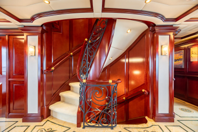 A luxurious wooden staircase with ornate railings is the focal point of the foreground, while the background reveals a hallway with artwork and patterned flooring.