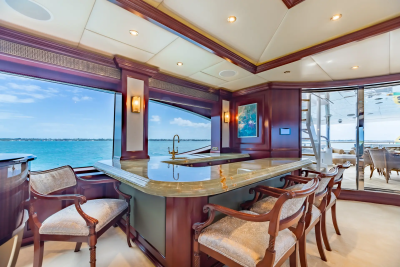 A luxurious yacht interior features a polished marble bar with upholstered stools in the foreground, while large windows reveal a bright blue ocean and sky in the background.