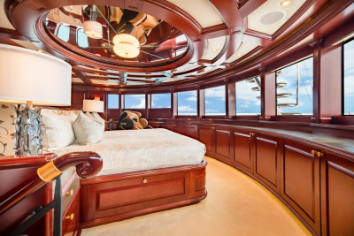 A luxurious bedroom with a plush bed in the foreground is surrounded by large windows offering a view of the ocean and a yacht in the background.