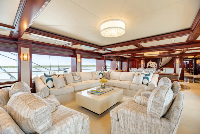 Plush armchairs and a sectional sofa with decorative pillows surround a cream-colored coffee table in the foreground, while a spacious dining area and a grand staircase are visible in the background through large windows overlooking the water.