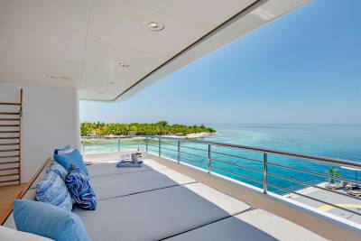 A luxurious yacht deck with plush loungers in the foreground overlooks a tropical island and turquoise ocean in the background.