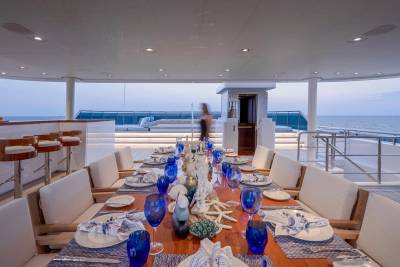 A long dining table set with blue glasses and white napkins is arranged on a yacht's deck, with the ocean and a hazy horizon visible in the background.