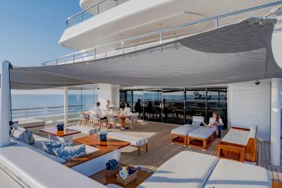 People relax on lounge chairs and at a dining table on a yacht's deck, with the ocean visible in the background.