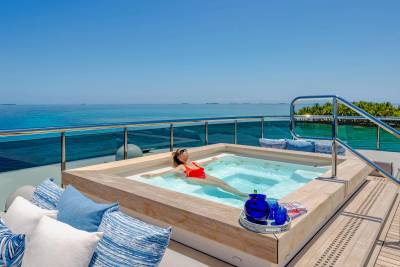 A woman relaxes in a hot tub on a deck with blue cushions, overlooking a calm turquoise ocean and distant islands under a clear blue sky.