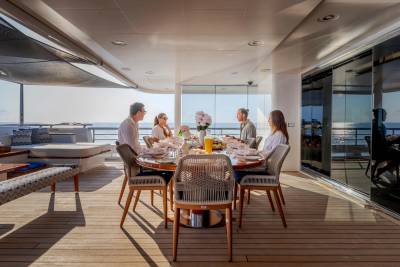 A group of people enjoy a meal at a round table on a yacht's deck, with the ocean visible in the background.