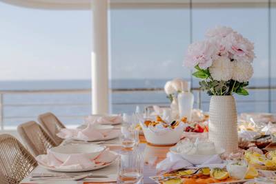 A beautifully set outdoor dining table with pink floral arrangements and a spread of food is positioned before a serene ocean backdrop.