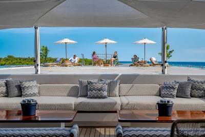 In the foreground, a plush sectional sofa with patterned pillows faces two wooden tables, while in the background, people relax under umbrellas on lounge chairs by the ocean.