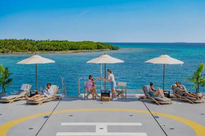 People relax on lounge chairs under umbrellas on a helipad with a tropical island and turquoise ocean in the background.