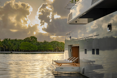 Sunlight glints off the sleek hull of a yacht with lounge chairs on its deck, while a lush, tropical coastline with palm trees lines the calm water under a dramatic, cloudy sky.