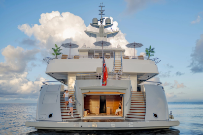 A couple walks up the stairs of a large yacht with an open garage door, while fluffy clouds drift in the blue sky behind.