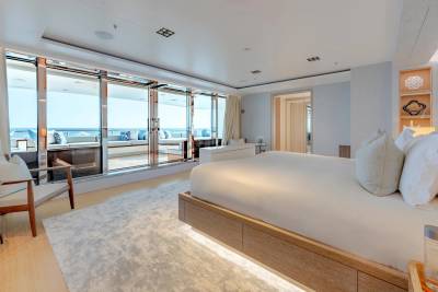 A luxurious bedroom with a large bed in the foreground opens onto a sunlit balcony with ocean views in the background.