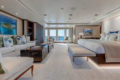 A luxurious bedroom with a plush white carpet, a large bed, and a seating area in the foreground, opens to a bright view of the ocean through large windows in the background.