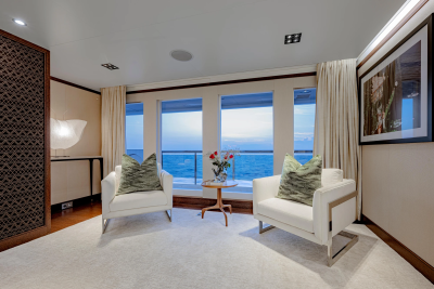 Two white armchairs with patterned pillows face a large window overlooking the ocean, with a small table between them and a framed artwork on the wall.