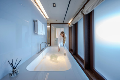 A woman in a white robe tests the water in a luxurious bathtub in a modern bathroom with large windows overlooking a bright, hazy exterior.