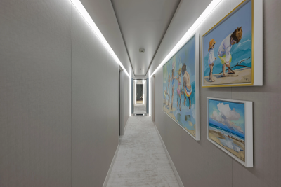 A long, narrow hallway with textured gray walls and a carpeted floor is illuminated by overhead lights, while colorful paintings of beach scenes adorn the right wall.